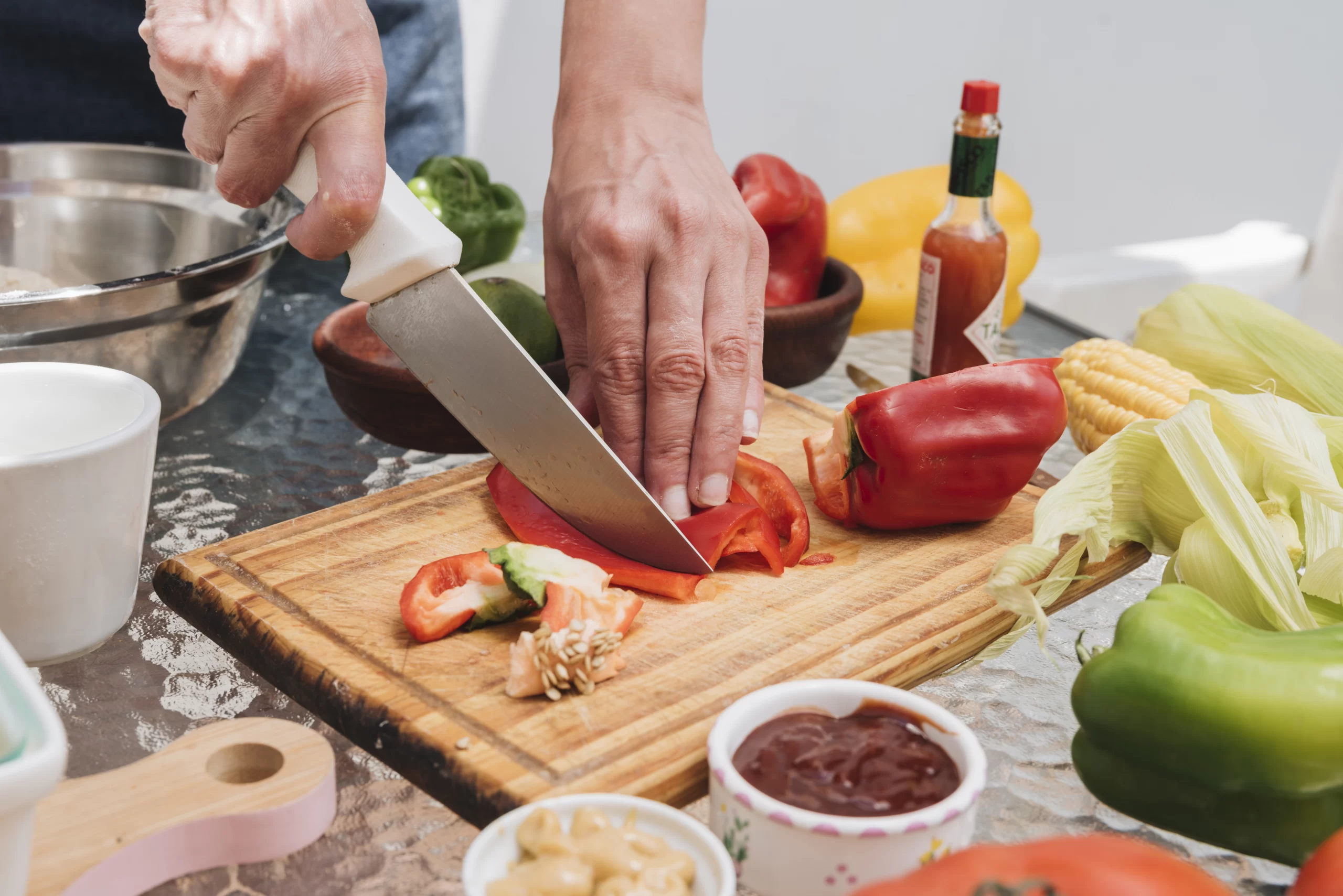 Person slicing red bell pepper on a cutting board surrounded by fresh vegetables and cooking ingredients in kitchen